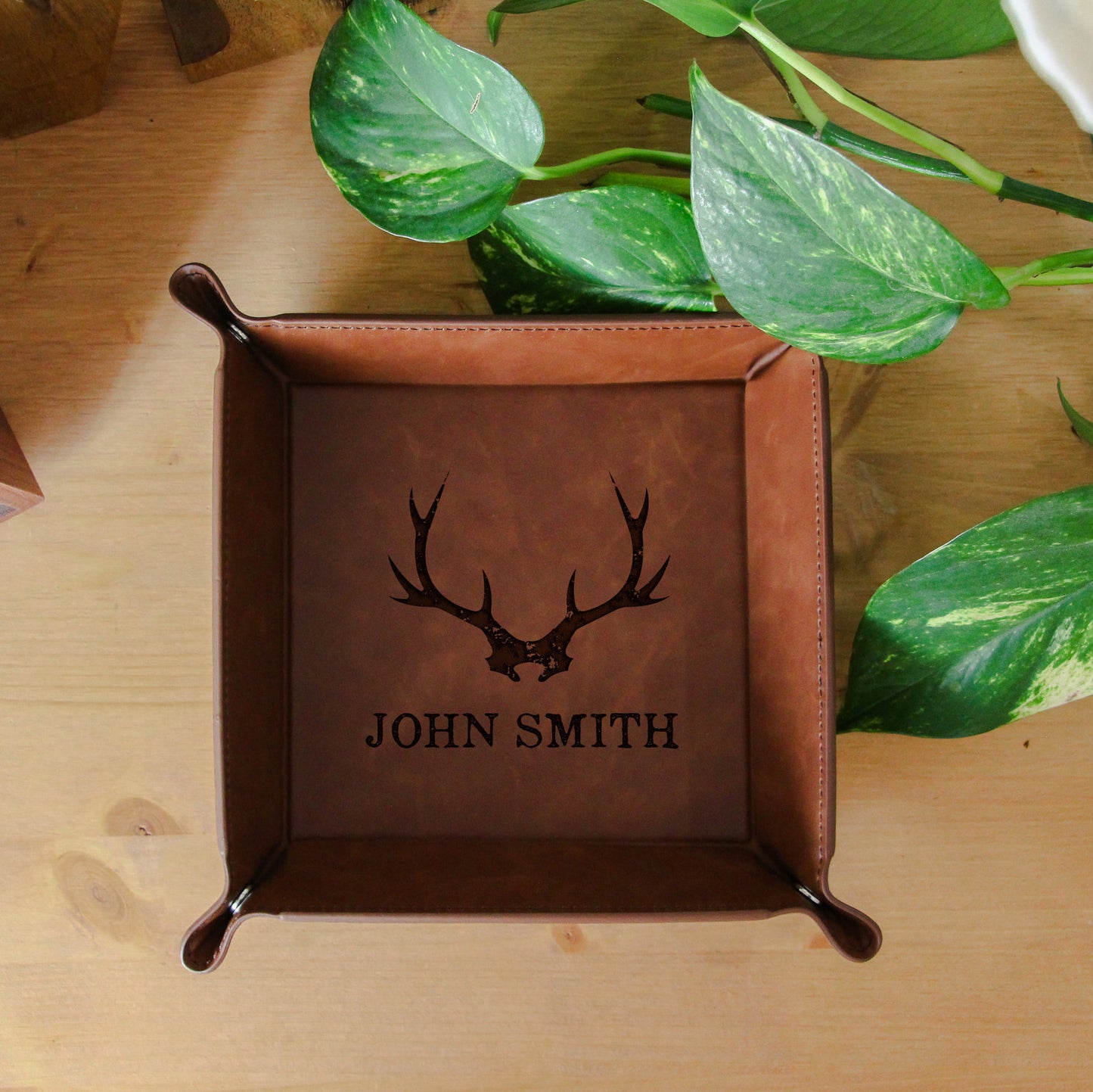 Brown leather tray with deer antlers and 'John Smith' engraving on a wooden surface with plants.
