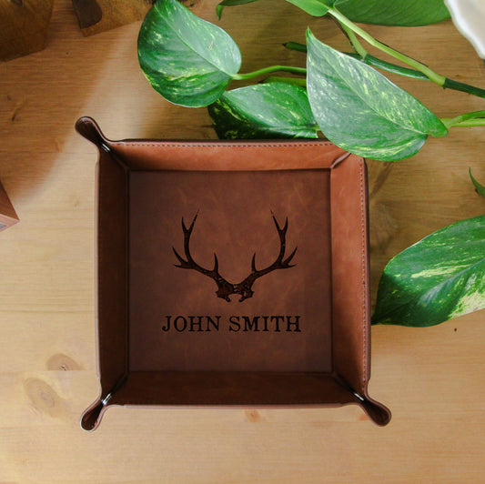 Brown leather tray with deer antlers and 'John Smith' engraving on a wooden surface with plants.